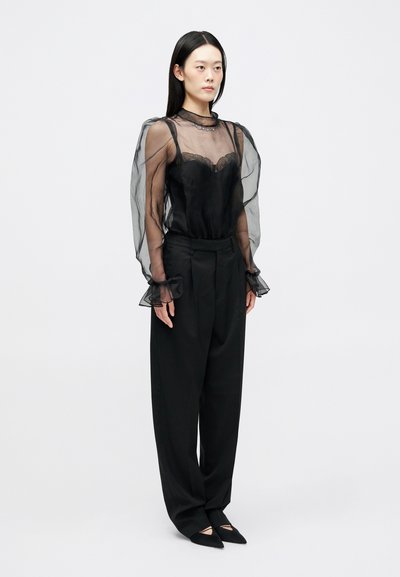 Μαύρη διάφανη μπλούζα με ρυτίδες στα μανίκια και δαντελένια λεπτομέ�ρεια, συνδυασμένη με φαρδιά μαύρα παντελόνια και μυτερές γόβες. Μινιμαλιστικός σχεδιασμός.