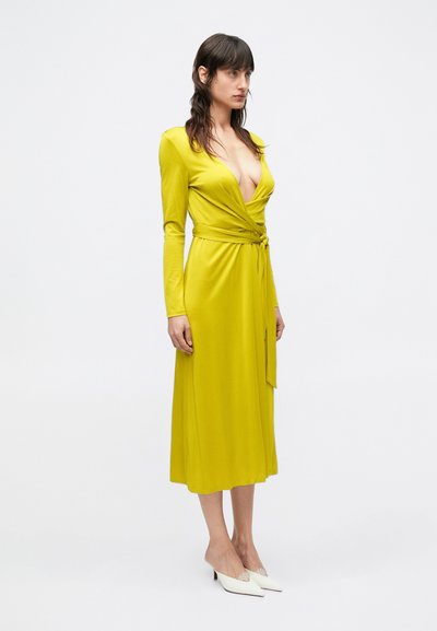 Rochie tip wrap galbenă dintr-un material ușor, cu mâneci lungi, decolteu adânc în V și talie cu cordon. Modelul a asortat-o cu pantofi cu toc și vârf ascuțit, albi.