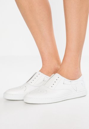 Bare legs wearing slip-on white leather sneakers with perforated decorative patterns and white rubber soles on a white background.