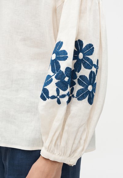 Κρεμ μακρυμάνικο με μπλε κέντημα λουλουδιών στη μανσέτα του μανικιού και συγκεντρωμένη, κουμπωτή μανσέτα, φορεμένο με σκούρο μπλε παντελόνι.