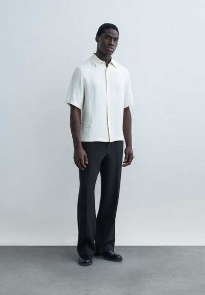 Άνδρας στέκεται μπροστά σε απλή τοίχο, φοράει κοντομάνικο κρεμ πουκάμισο, μαύρο παντελόνι και μαύρα παπούτσια, φαίνεται χαλαρός με τα χέρια στα πλευρά.