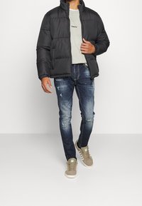 Giacca a vento nera con colletto alto; felpa grigia chiaro sotto; jeans blu strappati; sneakers beige con dettagli bianchi.