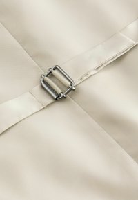 Beige Stoff mit glatter Textur, ordentlich genähten Nähten und einem silbernen Metallschnallen-Detail in der Mitte.