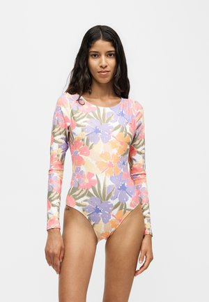 Ung kvinde med langt mørkt hår iført en heldragt med blomsterprint i pastel farver mod en ensfarvet lys baggrund.
