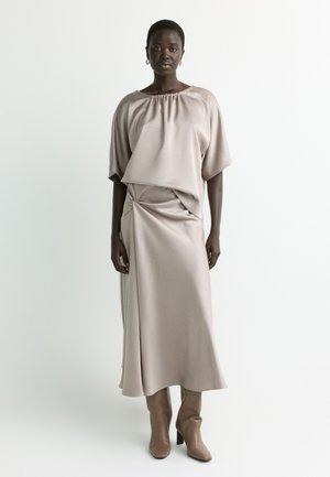 Dark-skinned woman wearing loose beige top, matching long skirt with draped knot, and tan heeled boots, standing against plain light background.