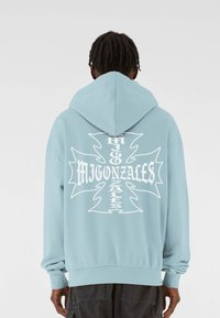 Svetlomodrá mikina s kapucňou s veľk�ým bielym grafickým potlačou, ktorá zobrazuje text a dizajn kríža na zadnej strane. Mäkko prístupný štýl s pretiahnutím.