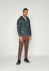 Μαύρη ζακέτα με κουκούλα, σε σκούρο πράσινο χρώμα, με υφή, συνδυασμένη με καφέ tapered παντελόνι. Συνδυάζεται με μαύρα αθλητικά παπούτσια.