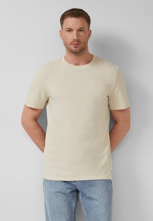 Man with short hair and beard wearing a textured cream short-sleeve shirt and light blue jeans, standing against a plain gray wall.