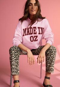 Růžová mikina s nápisem "MADE IN OZ" v barvě burgundské, spárovaná s džíny s leopardím vzorem. Model sedí na stříbrné židli před růžovým pozadím.