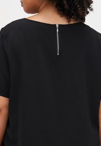 Top negro de manga corta con una tela texturizada y un cierre con cremallera centrada en la parte posterior. Cuello redondo simple y corte relajado.