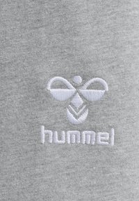 Hvidt broderet hummel-logo med stiliseret humlebi og tekst på lysegråt strikket stof.