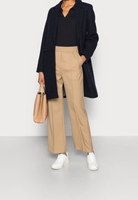 Marineblauer Mantel über schwarzem Oberteil, beigen weitem Hosen, und weißen Sneakern. Trägt eine hellbeige Handtasche. Minimalistisches Design, glatte Texturen.