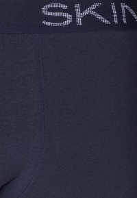 Marineblaue Leggings mit einer glatten Textur und einem elastischen Bund, der das Wort "SKIN" in Weiß zeigt und einen sanften Glanz reflektiert.
