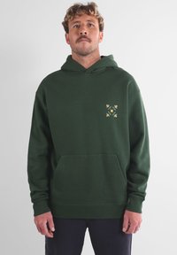 Sweat à capuche vert foncé avec une poche avant et un motif floral jaune et vert sur le côté gauche de la poitrine. Tissu doux et texturé.