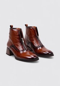 Botas de tobillo de charol marrón con punta afilada, cremallera frontal y tacón grueso de madera. Textura suave con un brillo sutil.