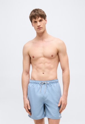 Young man with short hair wearing light blue swim shorts with a white drawstring, standing against a plain light background.