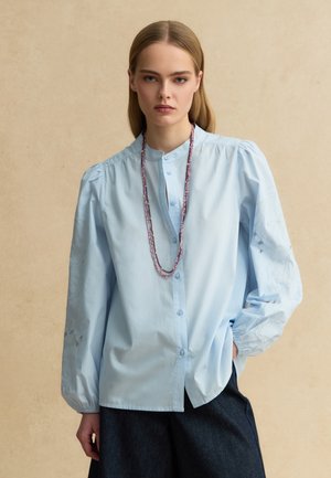 Junge Frau mit glattem Haar trägt eine hellblaue Bluse mit Puffärmeln und Knöpfen sowie eine lange Perlenkette, die Hände in den Taschen.