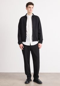 Schwarze Zip-Jacke über einem weißen Hemd, kombiniert mit schwarzen Hosen und Schuhen. Einfaches Design, glatte Textur und minimale Details.