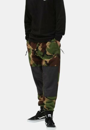 Pantalones jogger de camuflaje en forro polar verde y marrón con acentos de tela negra, que cuentan con una cinturilla elástica y puños en los tobillos, combinados con zapatillas negras.