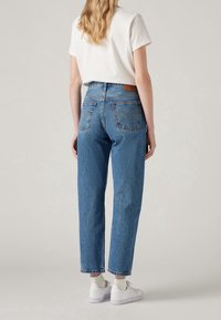 Vrouw met golvend blond haar, gekleed in een wit T-shirt, hooggeknoopte blauwe jeans, witte sokken en witte sneakers, staand tegen een effen achtergrond.