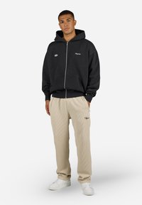 Sort hættejakke med lynlås, fører lommer og broderet logo, parret med beige ribbede bukser og hvide sneakers. Simpelt, afslappet outfit.