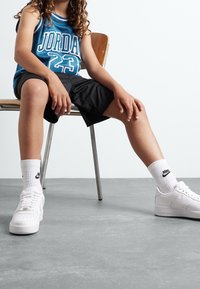 Modrý síťovaný basketbalový dres s nápisem „JORDAN 23“, černé šortky, bílé ponožky Nike a bílé tenisky Nike Air Force 1. Sedí na židli.