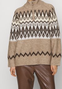 Maglione knit beige con colletto a mezza zip, caratterizzato da motivi geometrici in marrone scuro e crema. Tessuto morbido, vestibilità ampia e polsini a coste.