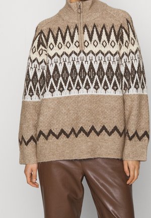 Beige gebreide trui met een halve ritssluiting, met geometrische patronen in donkerbruin en crème. Zachte textuur, losse pasvorm en geribde manchetten.