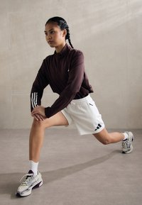 Bordo ilgomis rankovėmis marškiniai su baltais juostelėmis, baltos šortai su juodu Adidas logotipu ir daugiaspalvės sportinės bateliai, tempiasi lunge.