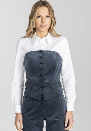 Granatowa kamizelka z korduroy o dopasowanym kroju, z przodem zapinanym na guziki i bocznymi kieszeniami, w połączeniu z białą koszulą z kołnierzykiem pod spodem.