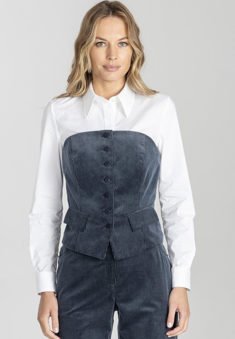 Granatowa kamizelka z korduroy o dopasowanym kroju, z przodem zapinanym na guziki i bocznymi kieszeniami, w połączeniu z białą koszulą z kołnierzykiem pod spodem.