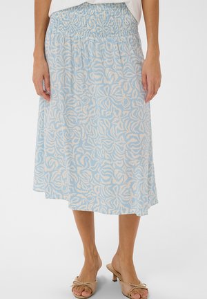 Kobieta w jasnoniebieskiej spódnicy do kolan z białymi, abstrakcyjnymi wzorami kwiatowymi i marszczoną talią, zestawiona z beżowymi sandałami na obcasie z odkrytymi palcami.