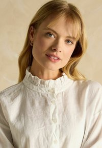 White linen blouse featuring a ruffled collar, button-down front, and long sleeves. Textured fabric with a light, airy appearance.