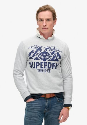 Sweatshirt gris avec un graphique de montagne bleu marine, texte "SUPERDRY" et poignets côtelés, associé à un jean bleu et une ceinture marron.