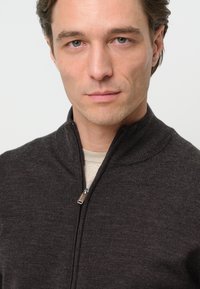 Sweater de cremallera de color gris oscuro con un cuello alto. Hecho de material de punto, presenta una textura suave y un brillo sutil. Detalle de cremallera plateada.