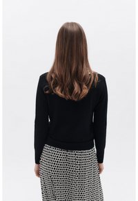 Maglione nero a maglia con maniche lunghe, abbinato a una gonna a fantasia con un motivo a catena in bianco e nero. Modella vista da dietro.