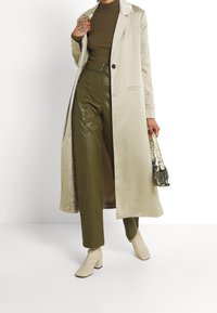 Manteau long en satin beige, pantalon en simili cuir vert olive avec une ceinture, bottes à talons bloc beiges et un petit sac à main en imprimé serpent noir et blanc.