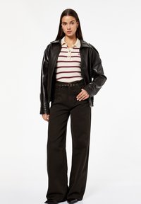 Donna con una giacca di pelle nera sopra una polo bianca a righe, pantaloni larghi neri con cintura e scarpe nere, in piedi contro uno sfondo bianco.