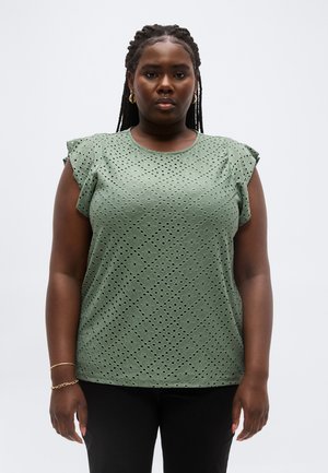 Kvinde med flettet hår iført guldfarvede øreringe i hoop-stil og grøn top med korte ærmer og øjesløjer, stående foran en ensartet lys baggrund.