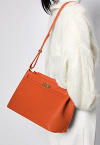 Orange läderhandväska med en strukturerad form, utrustad med ett guldhårdvarulås och en lång justerbar rem. Texturerat yttermaterial synligt.