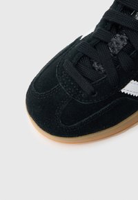 Črni semiš tekaški copat z teksturirano površino, z belimi črtami, vezalkami in gumijastim podplatom v barvi gume. Povečani pogled na predel prstov.