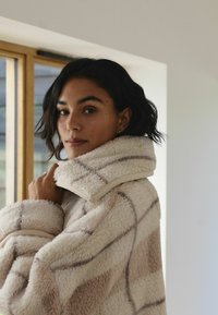 Kremowa, oversize'owa kurtka z miękkiego, strukturalnego polaru z wzorem w jasnobrązowym kolorze oraz wysokim kołnierzem.