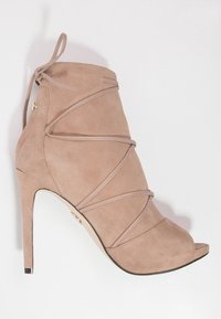 Beige ruskindsankelstøvle med peeptoe-design, krydsseler, høj stiletthæl og metallisk detalje nær lynlåslukning.
