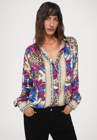 Kvinde med mørkt bølget hår iført en blomstret og leopardprint bluse med "Just Cavalli" tekst, hænder i lommen på sorte bukser, neutral baggrund.