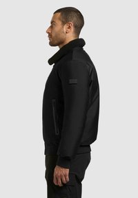 Svart bomberjakke med sherpa-krage, glidelåslommer på sidene og ribbestrikkede mansjetter. Har et merkebånd på venstre erme.