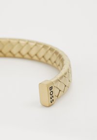 Brățară metalică împletită din aur, cu un capăt dreptunghiular neted pe care este inscripționat cuvântul "BOSS" în negru. Suprafață plană, design geometric și finisaj texturat.