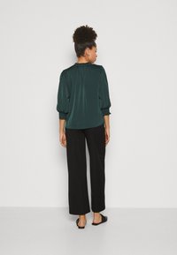 Blouse en satin vert foncé avec des manches bouffantes et des poignets élastiques, associée à un pantalon large noir et des ballerines noires, vue de dos.