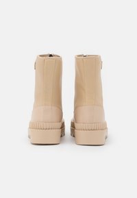 Un par de botas de tobillo beige con suelas gruesas, vistas desde atrás sobre un fondo blanco.