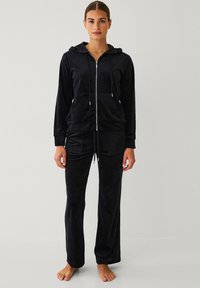 Ensemble de survêtement en polaire noire avec un sweat à capuche zippé et un pantalon à cordon de serrage. Il présente une texture lisse et des poches avant. Coupe décontractée, sans logos visibles.