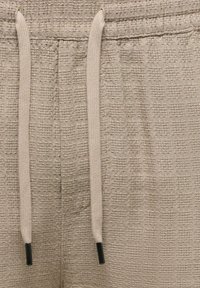 Tissu beige texturé avec un motif carré tissé et des cordons beiges avec embouts en plastique noir sur une ceinture de vêtement.